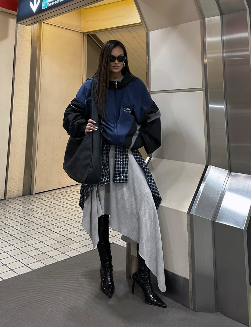Mulher em estação de metrô com jaqueta azul, botas pretas e saia assimétrica. Look reflete tendências vintage com um toque contemporâneo.