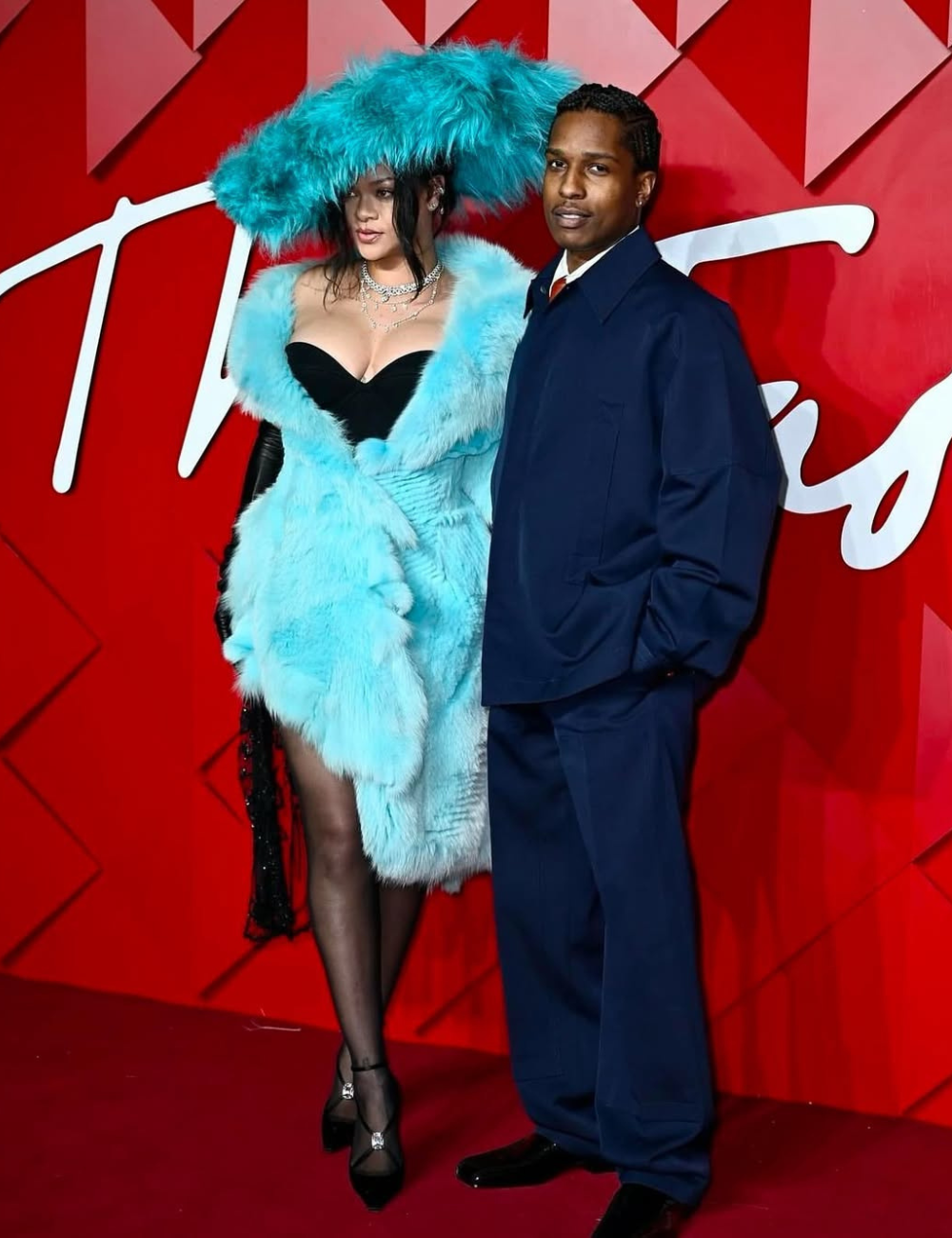 Vestindo um glamouroso casaco azul e chapéu combinando, a pessoa destaca as tendências vintage em um evento elegante, ao lado de outra figura em traje formal.