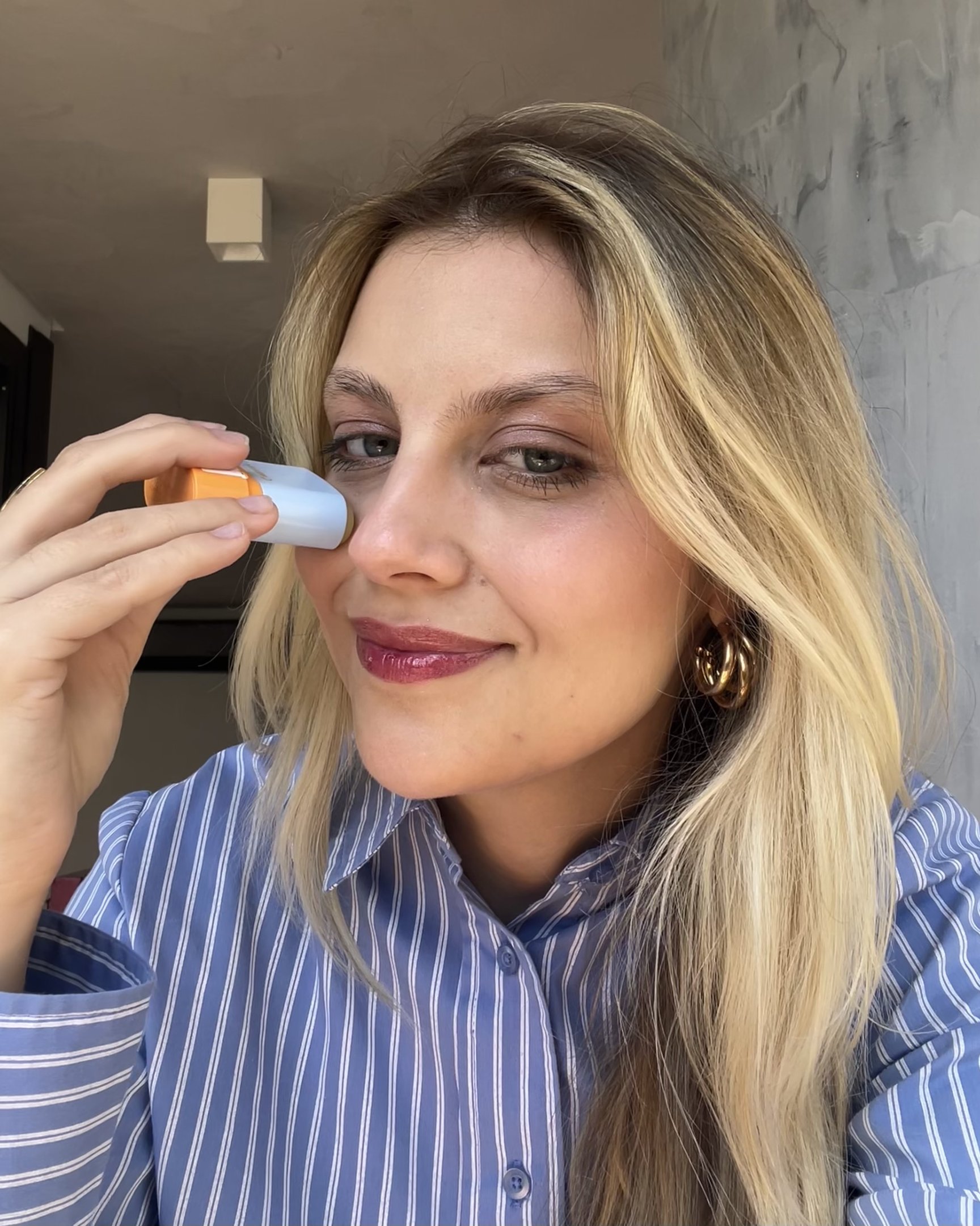 Mulher aplicando protetor facial em bastão com embalagem azul e laranja. Ela sorri levemente, vestindo camisa listrada.