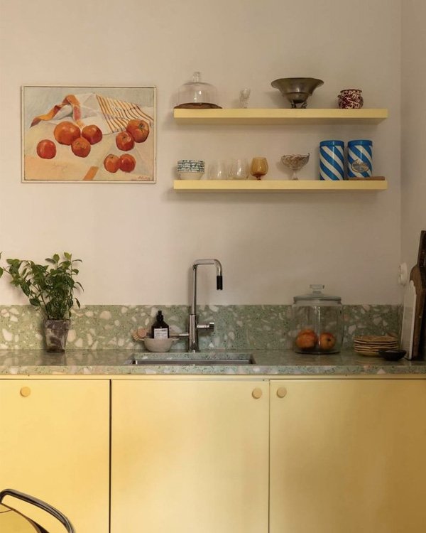 Cozinha com armários amarelos, prateleiras decoradas e um quadro de frutas, ajudando a decorar a casa com charme.