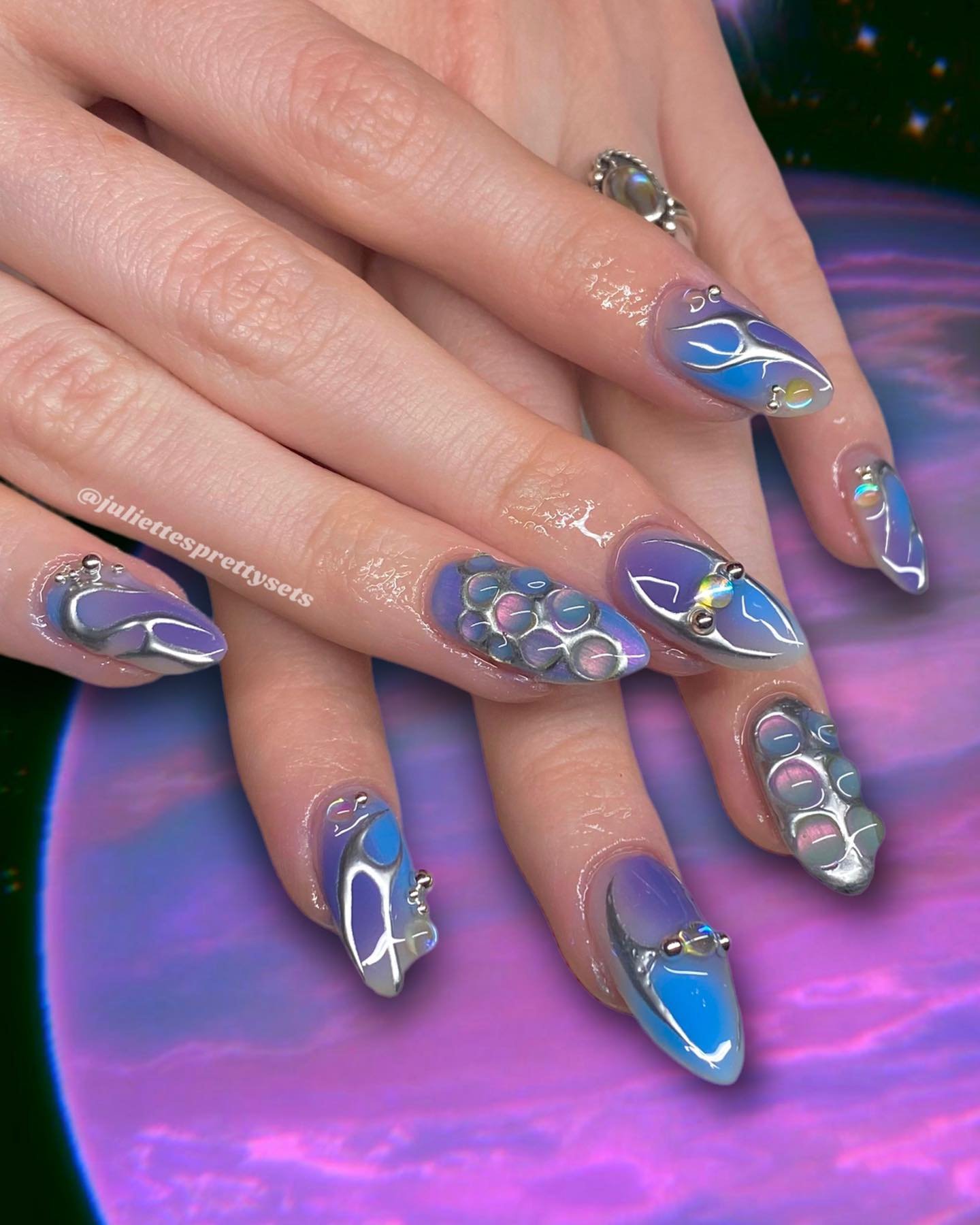 Unhas com design 3D em tons de roxo e azul, decoradas com detalhes metálicos e esferas brilhantes.