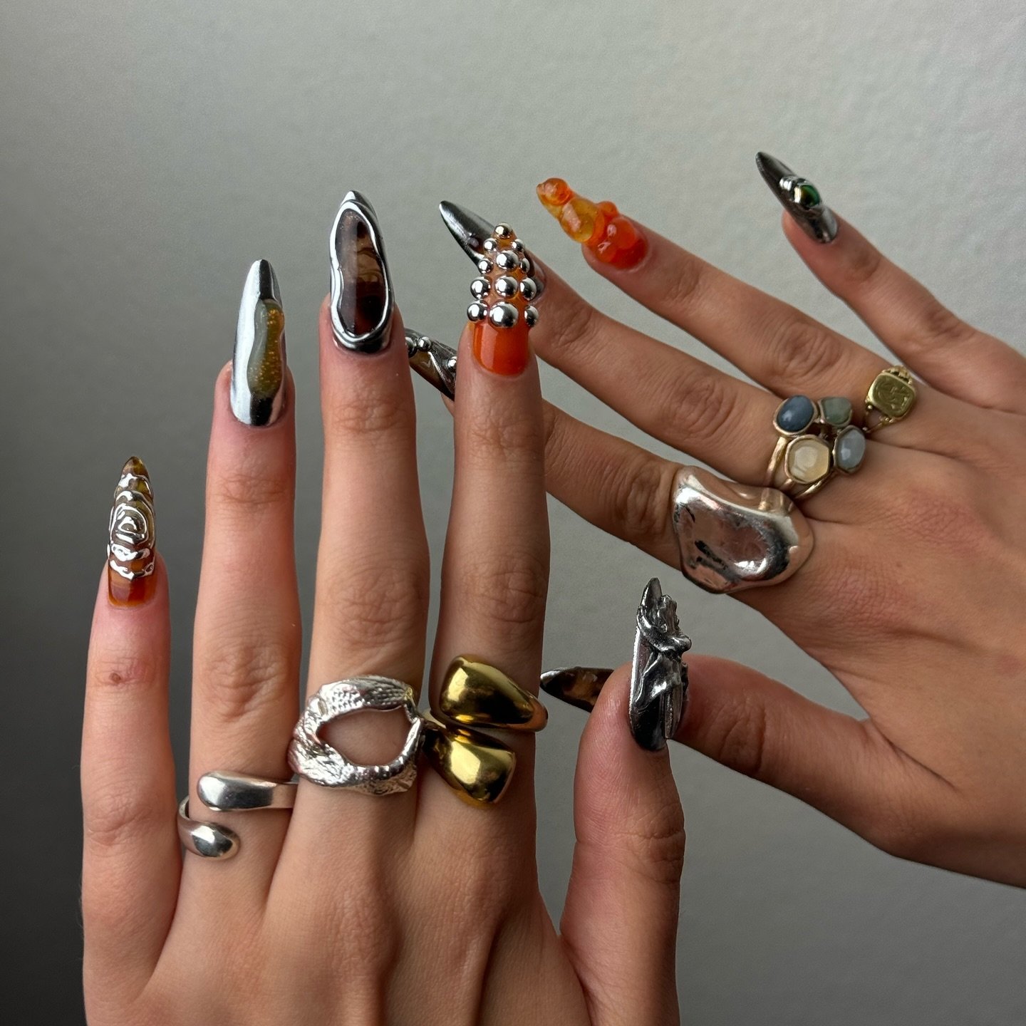 Unhas com design 3D metálico e laranja, adornadas com anéis variados e detalhes chamativos.