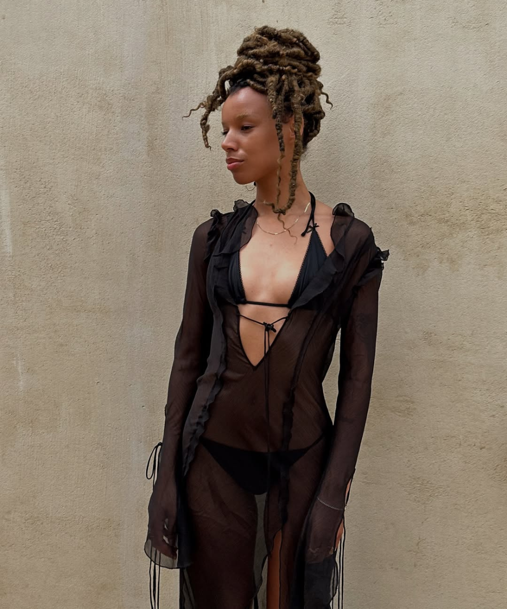 Modelo em vestido preto transparente e biquíni, estilo típico para um verão em Ibiza, em frente a uma parede neutra.