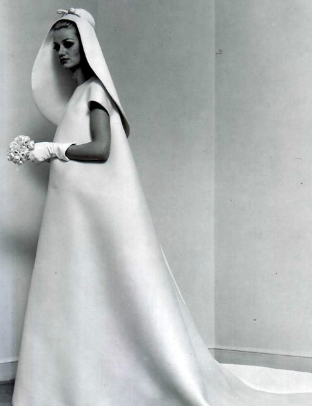 Modelo com vestido de noiva de alta-costura, capuz e luvas, segura pequeno buquê em foto vintage.