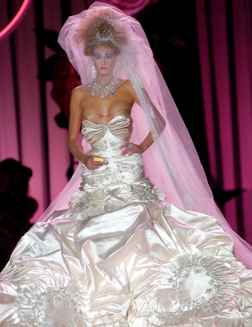 Modelo com vestido de noiva da alta-costura volumoso, tecido acetinado e detalhes brilhantes, desfilando com véu e tiara em passarela iluminada.