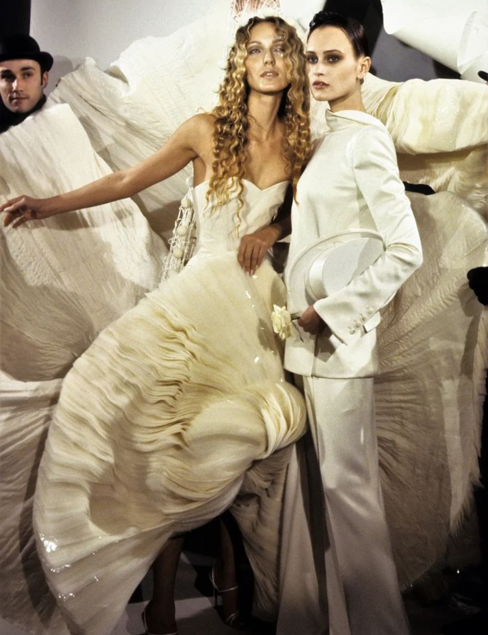 Modelos posam com vestidos de noiva da alta-costura; um em vestido volumoso e outro em terno elegante.