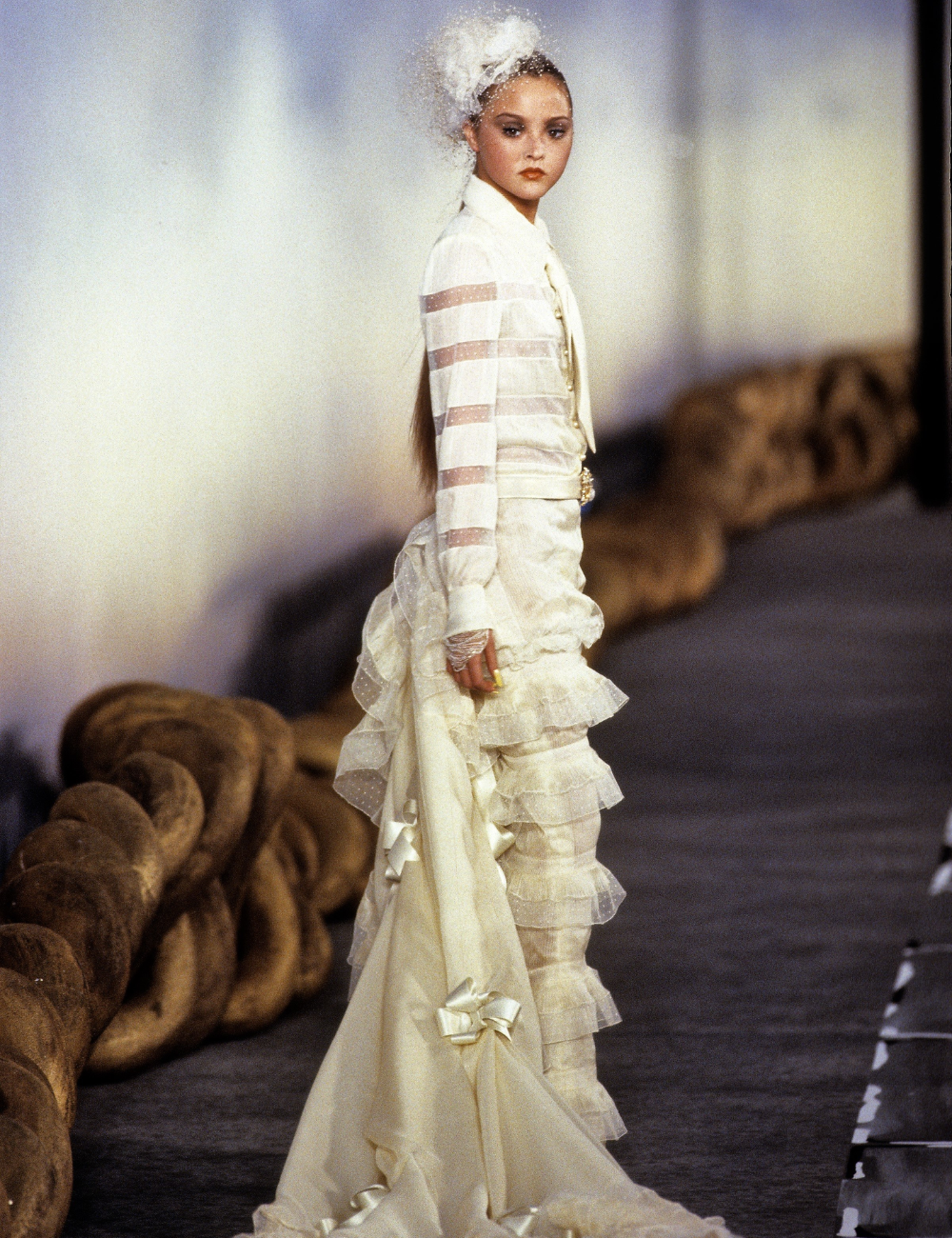 Modelo desfilando em vestido de noiva da alta-costura, com detalhes em camadas e cauda elegante.