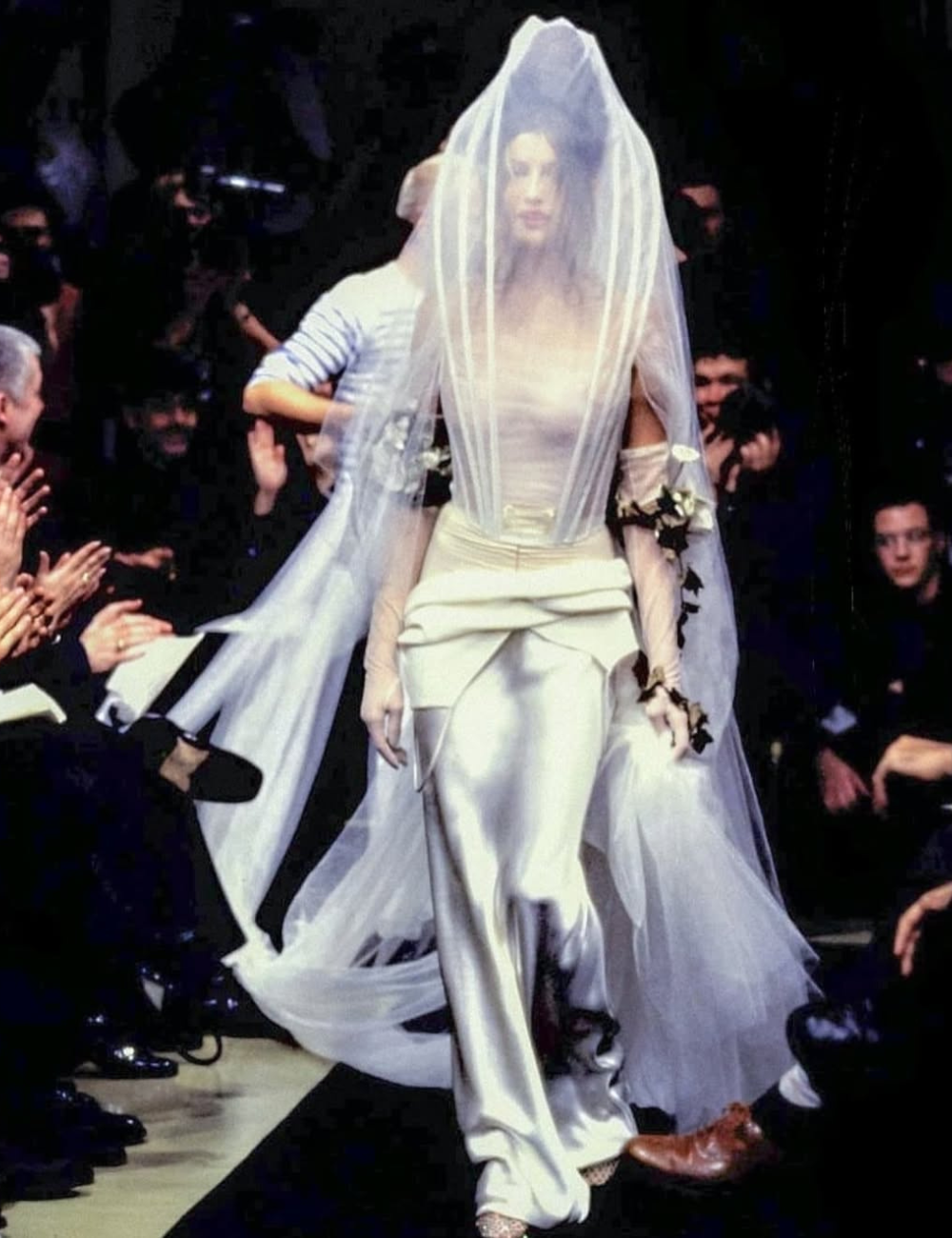 Modelo usando vestido branco elegante com véu longo em desfile de alta-costura. Público aplaude ao redor da passarela.