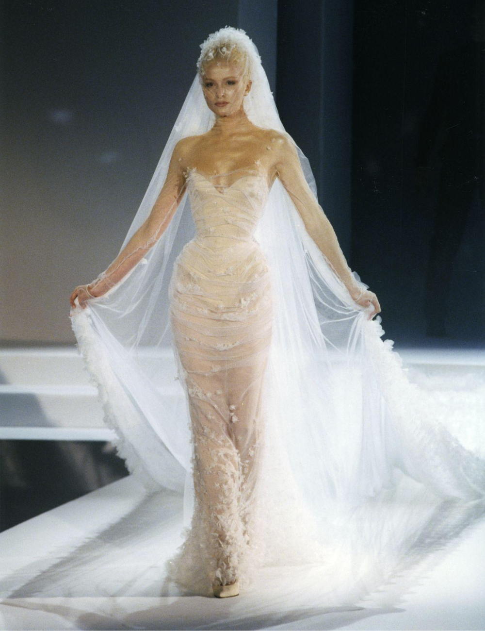 Modelo desfilando em vestido de noiva da alta-costura, com longo véu branco e detalhes delicados.