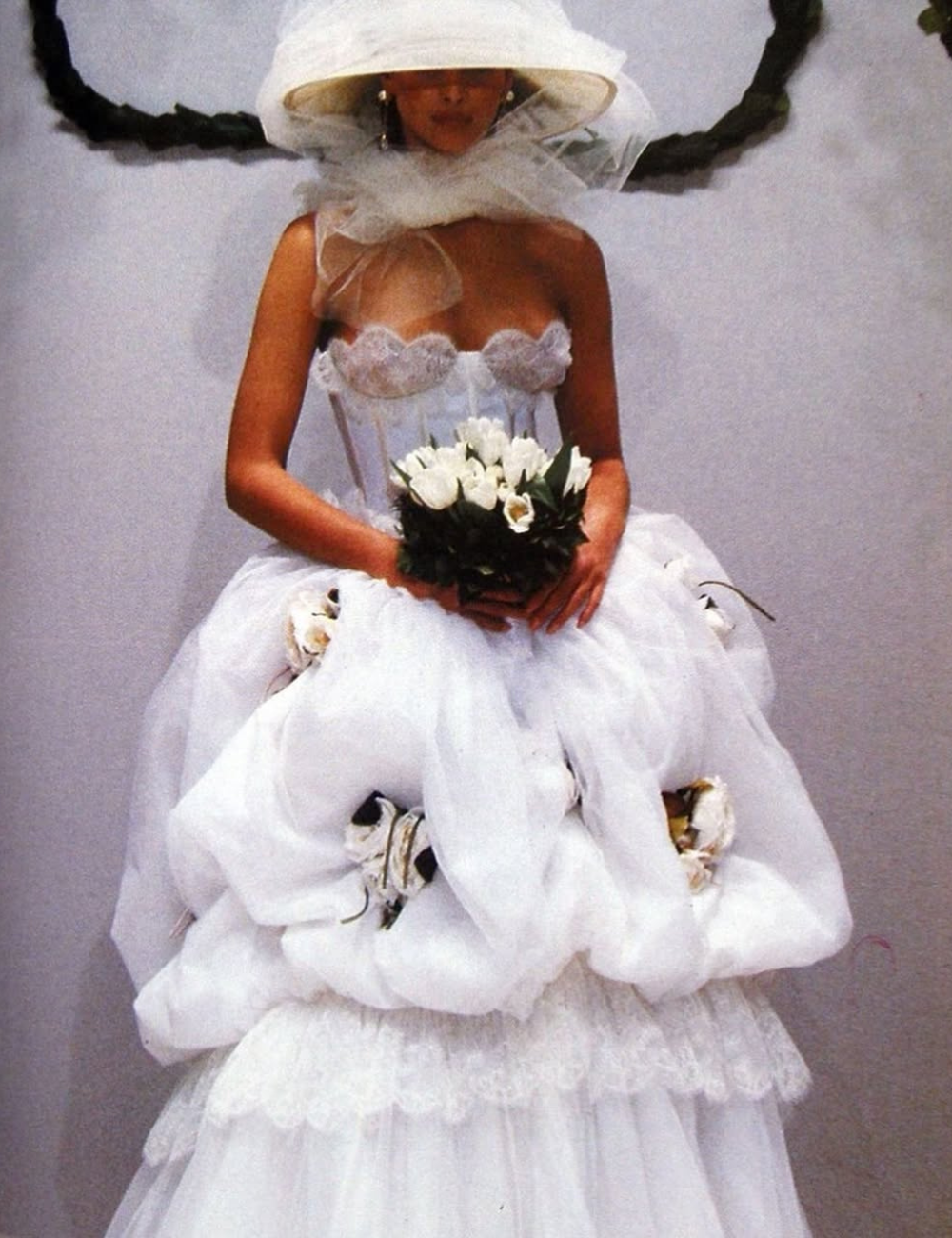 Noiva com vestido de alta-costura, repleto de camadas e flores brancas, além de chapéu e véu elaborados, segurando buquê.