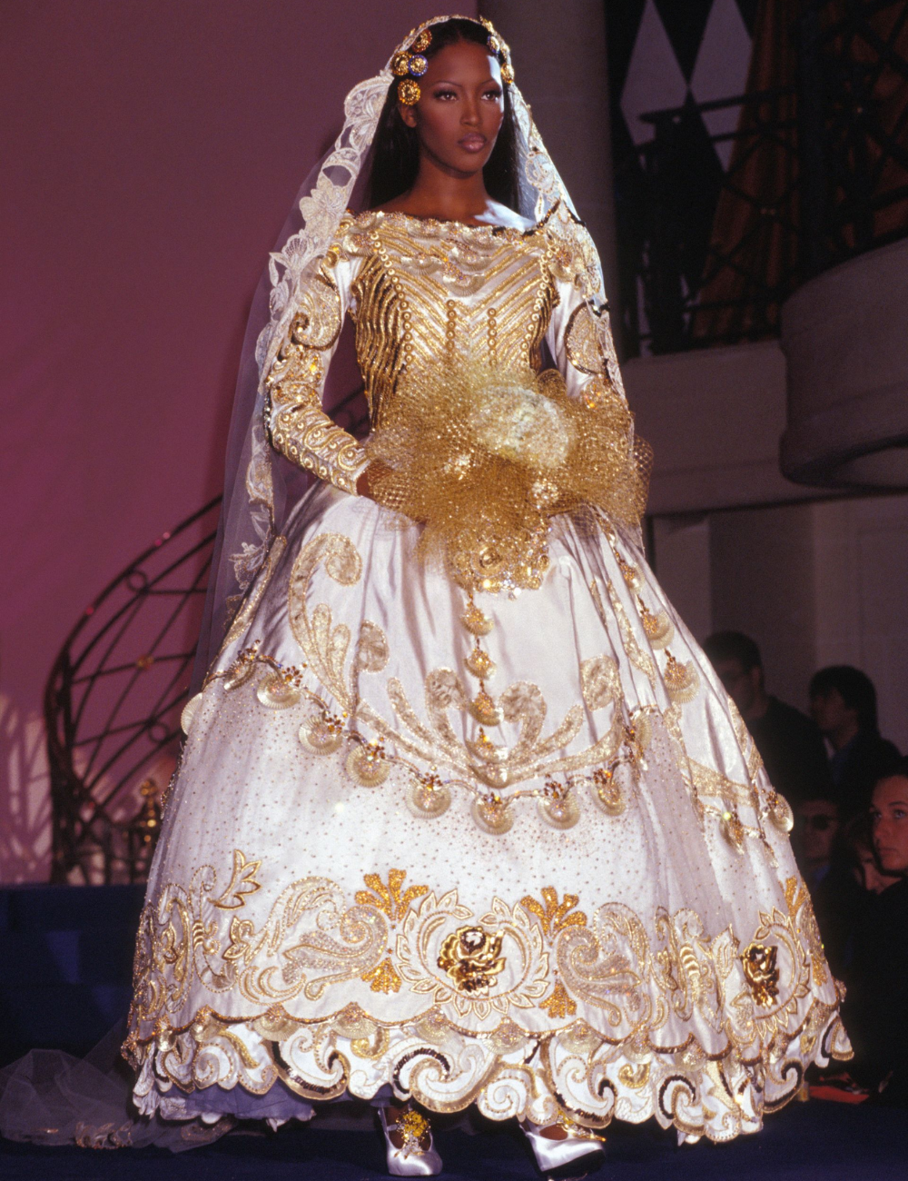 Modelo usando vestido de noiva da alta-costura em tons dourados e detalhes ornamentados, segurando um buquê dourado.