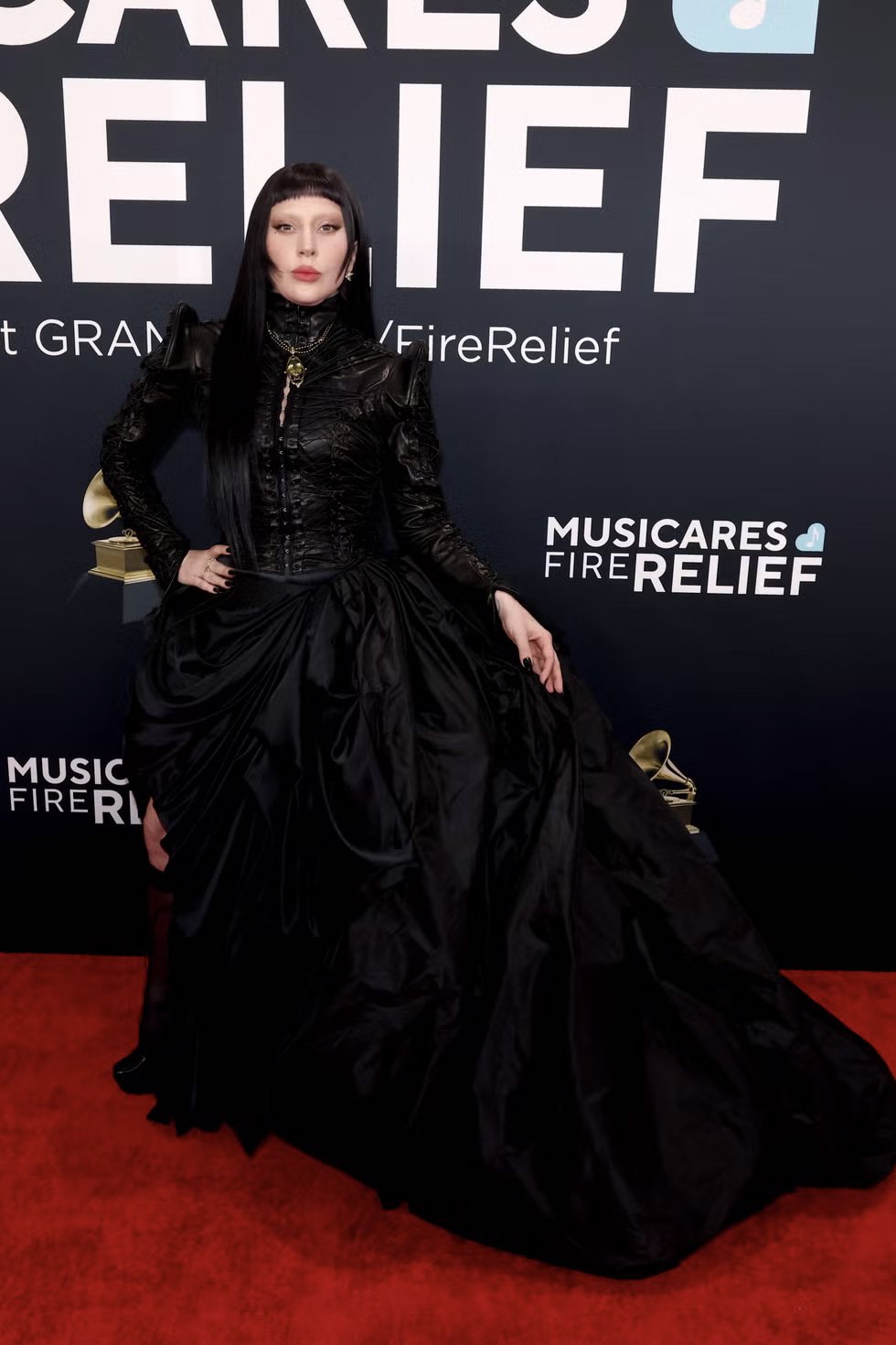 Lady Gaga de vestido preto elegante posando em tapete vermelho em evento beneficente.