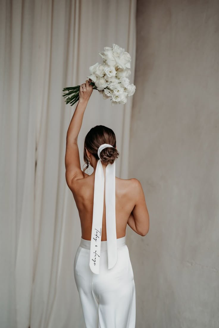Mulher de costas com cabelo preso por fita branca, segurando um buquê de flores, destacando acessórios de noiva.