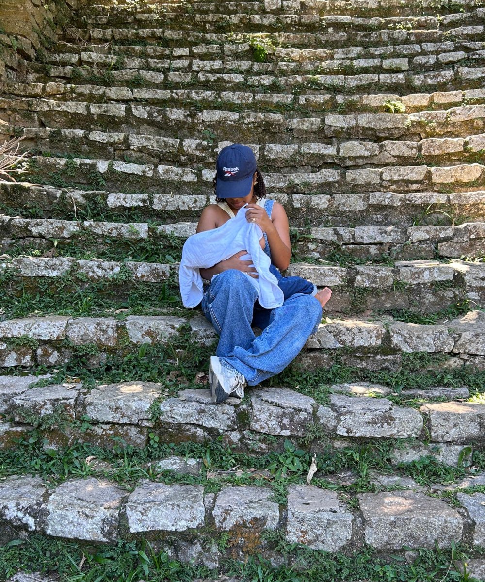 Pessoa sentada em degraus de pedra, cuidando de bebê coberto por pano branco. Bomba extratora