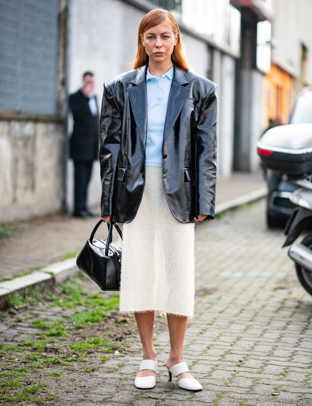Mulher usando casaco de couro preto sobre blusa azul, saia branca texturizada e sapatos elegantes. Peça estilosa completa o look.