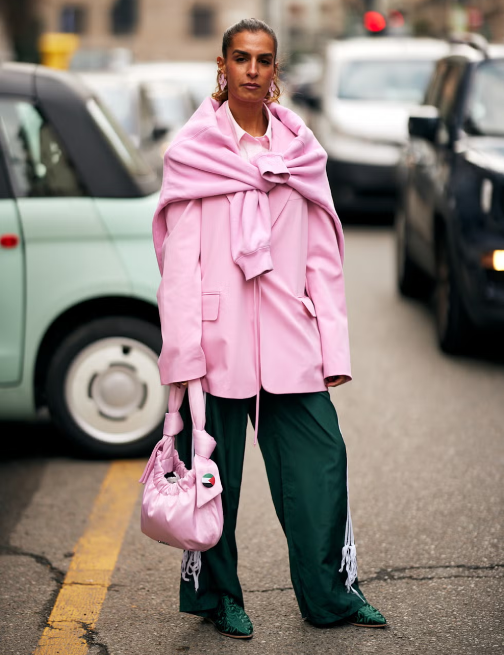 Pessoa vestindo blazer rosa, calças verdes e carregando bolsa combinando. Uma peça estilosa em meio ao trânsito urbano.