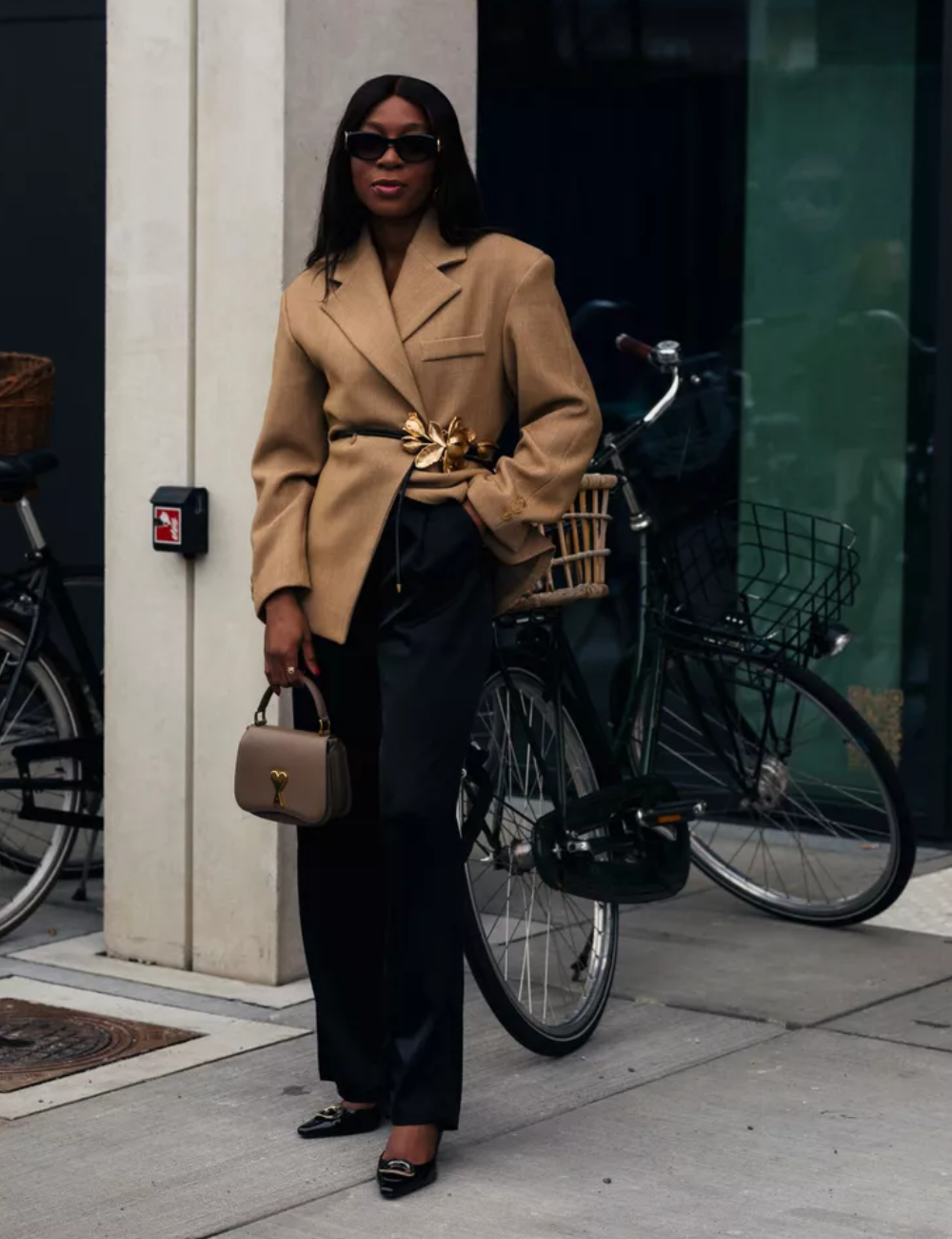 Mulher usa blazer bege estiloso com cinto dourado, calça preta e óculos de sol, carregando bolsa.