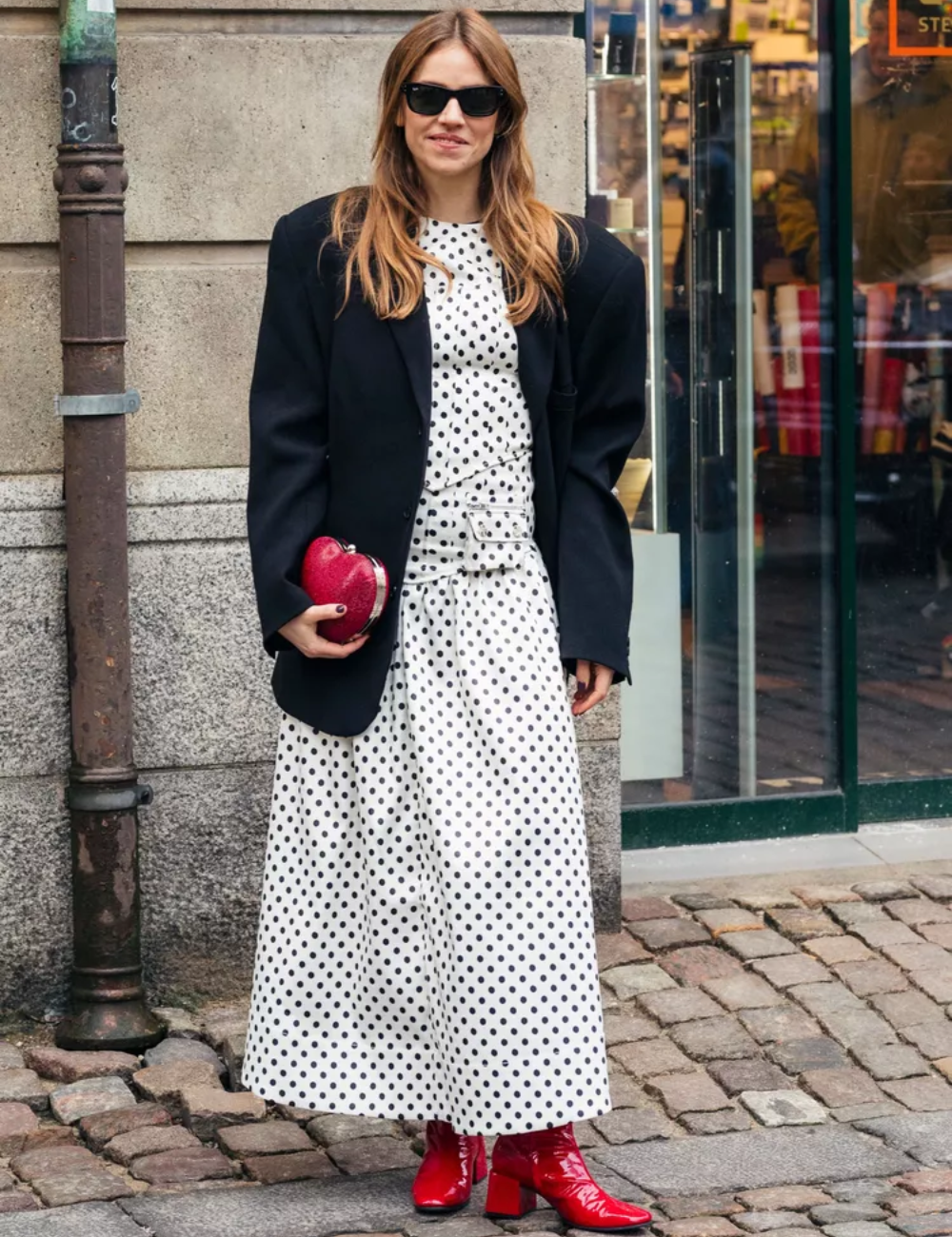 Pessoa com vestido de bolinhas preto e branco, blazer preto e botas vermelhas, representando uma peça estilosa.