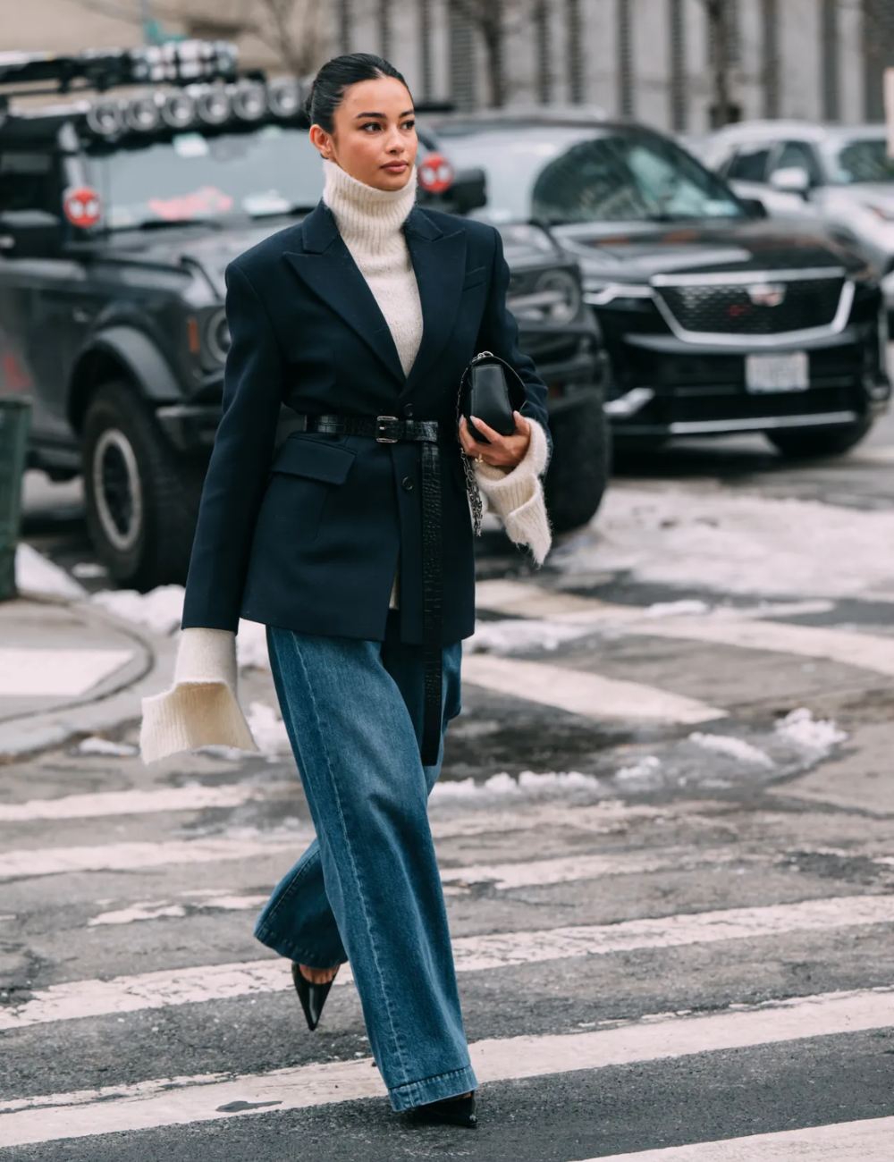 Mulher usando blazer preto, suéter de gola alta e calça jeans larga, exibindo uma peça estilosa enquanto caminha na cidade.