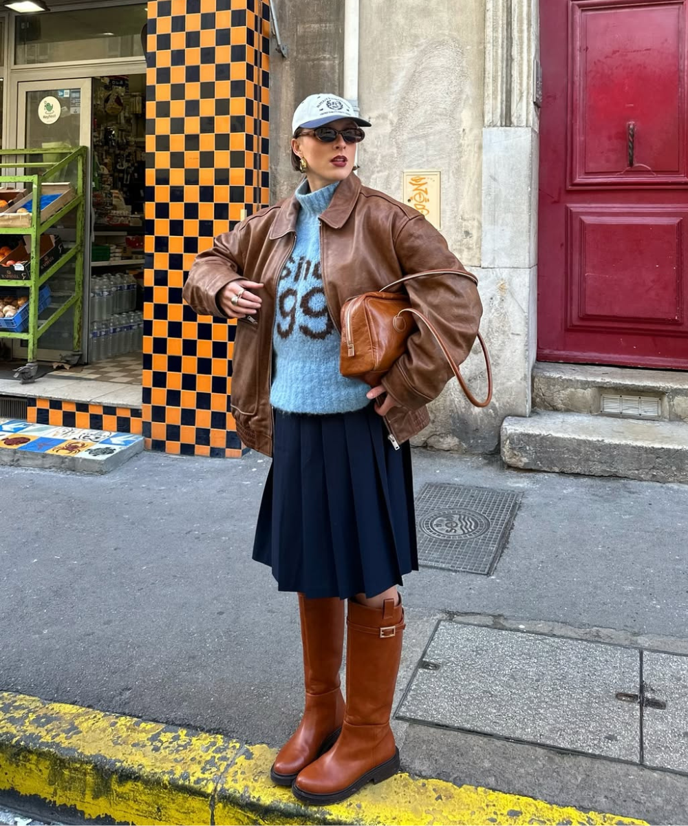 Pessoa usando suéter azul, jaqueta marrom e saia plissada preta com botas femininas 2025 modernas.