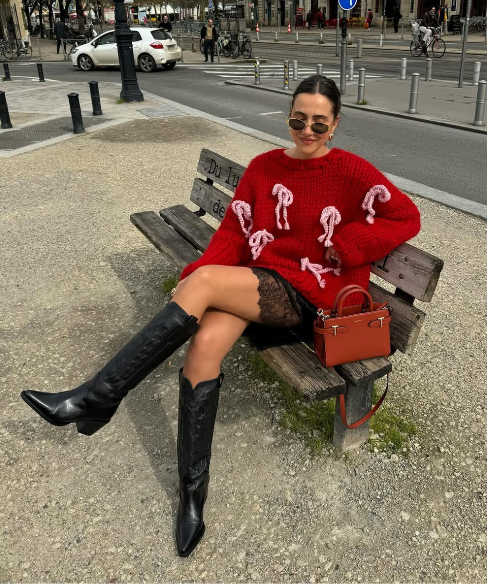 Mulher em banco com suéter vermelho, saia rendada preta e botas femininas destacando estilo 2025. Bolsa laranja ao lado.