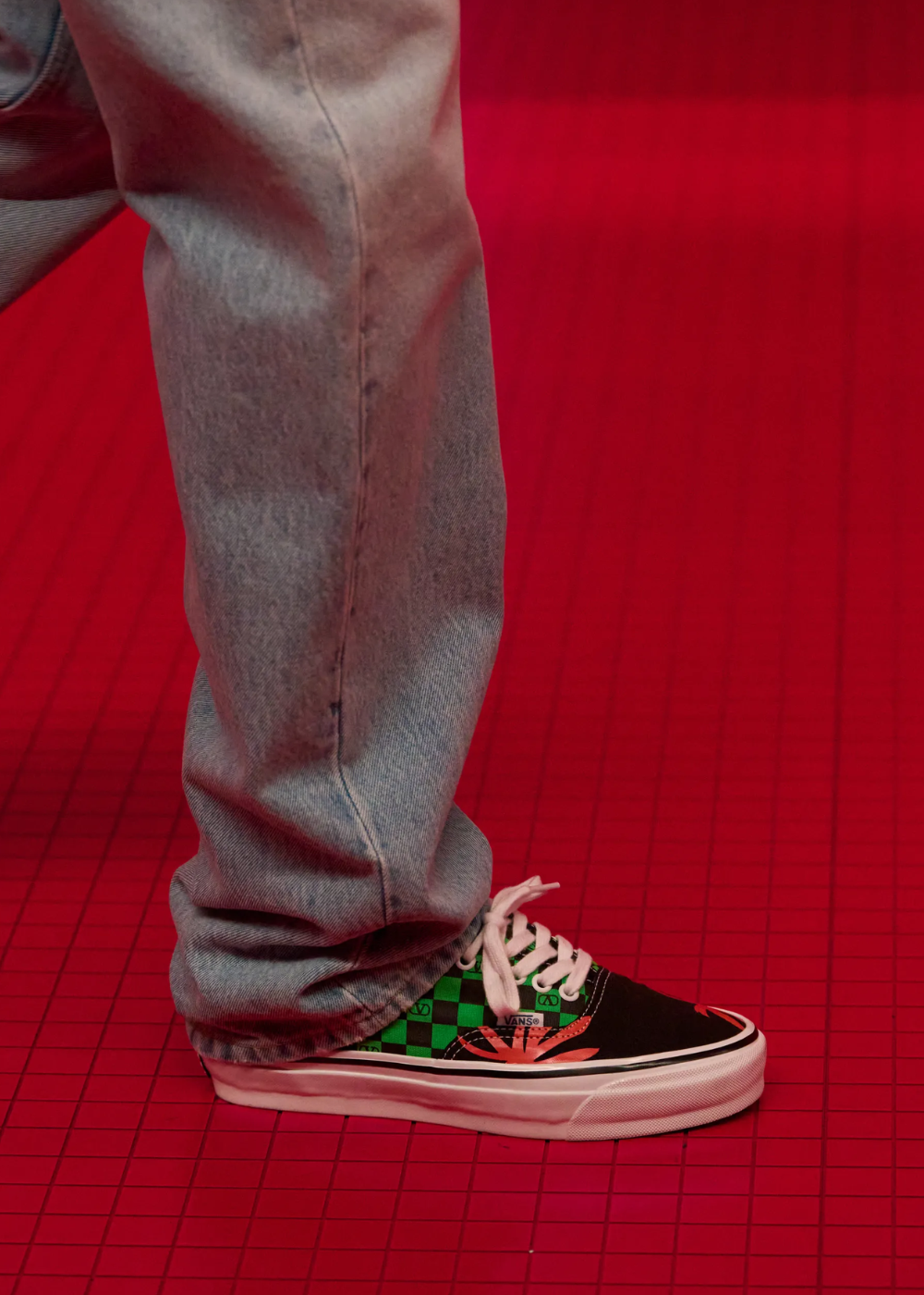 Tênis verde e preto com padrão xadrez, colaboração estilosa, usado com calça jeans em piso vermelho.