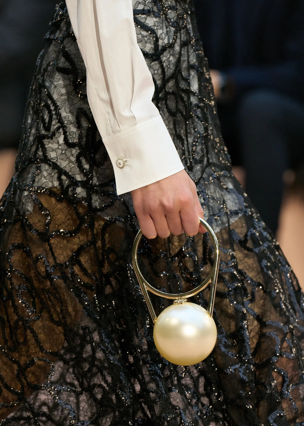 Mão segurando bolsa em forma de pérola com alça dourada, em contraste com saia rendada preta.