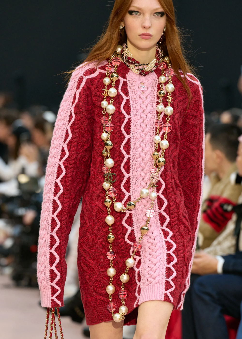 Modelo em desfile usando suéter vermelho e rosa com colar de pérolas longas, destacando o uso de joias.