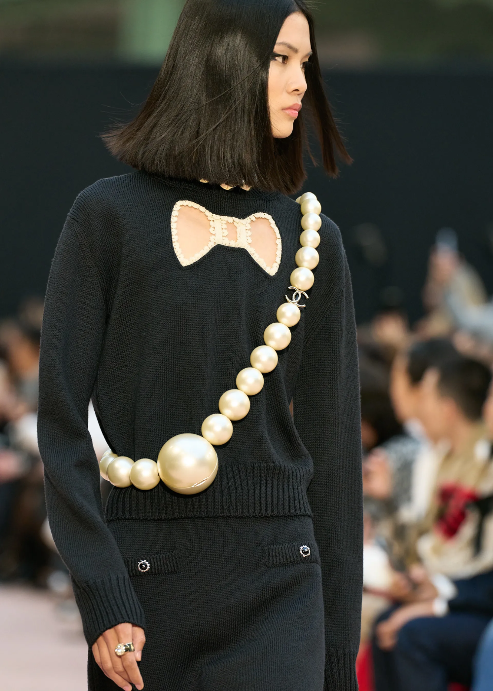 Desfile de moda com modelo usando suéter preto decorado com laço e pérolas grandes, explorando como usar pérolas.