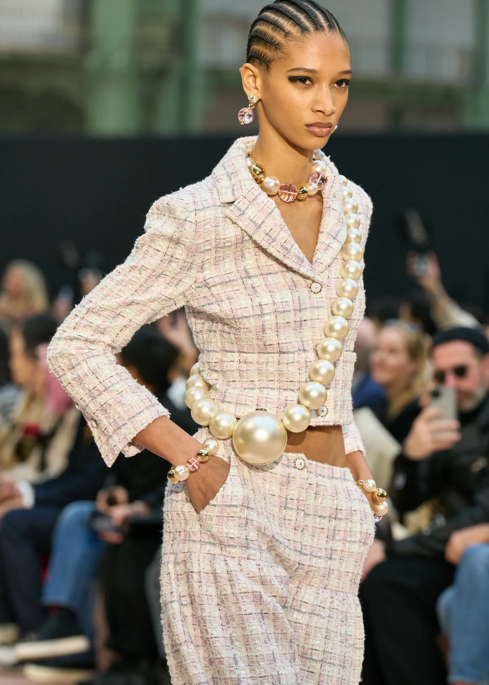 Modelo desfilando com colar e cinto de pérolas grandes, usando conjunto de tweed rosa e branco em uma passarela.