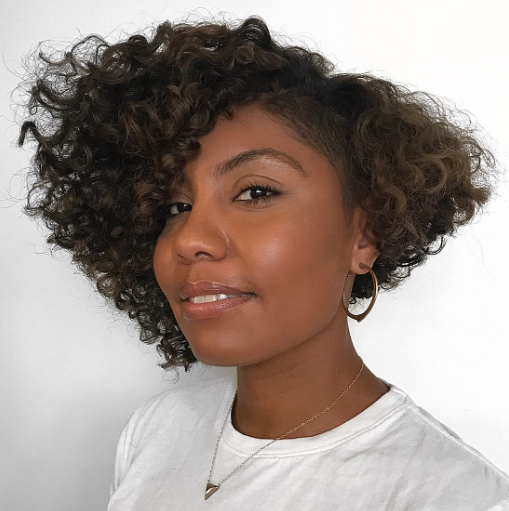 Mulher com cortes de cabelo crespo e cacheado volumoso em corte assimétrico, usando brincos de argola e camiseta branca.