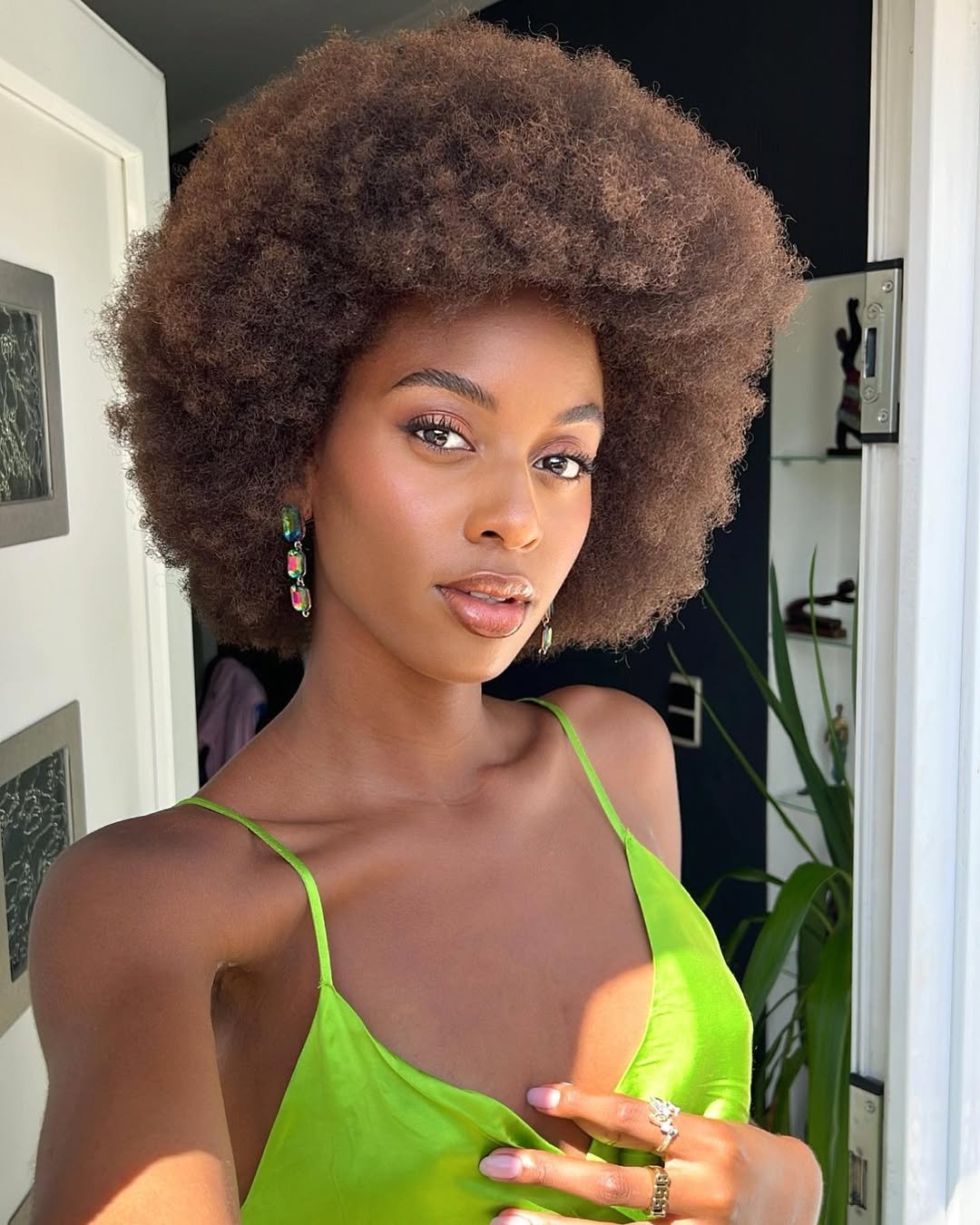 Mulher com cabelo afro volumoso usa vestido verde e brincos coloridos, representando cortes de cabelo crespo e cacheado.