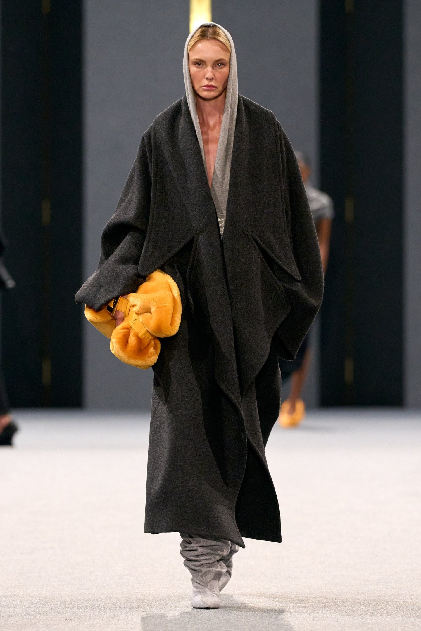 Modelo em manteaux cinza e botas na passarela do desfile da Balmain. Ela segura acessório laranja.