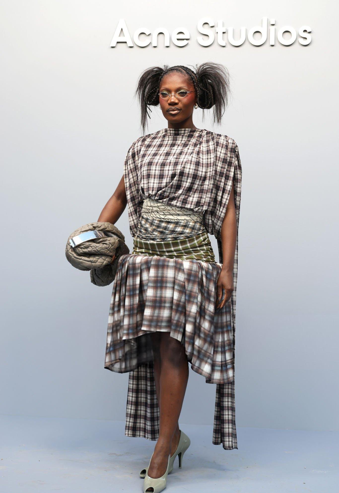 Pessoa em vestido xadrez assimétrico na frente de logotipo da Acne Studios, com cabelo estilizado em dois coques altos. Looks da Doechii