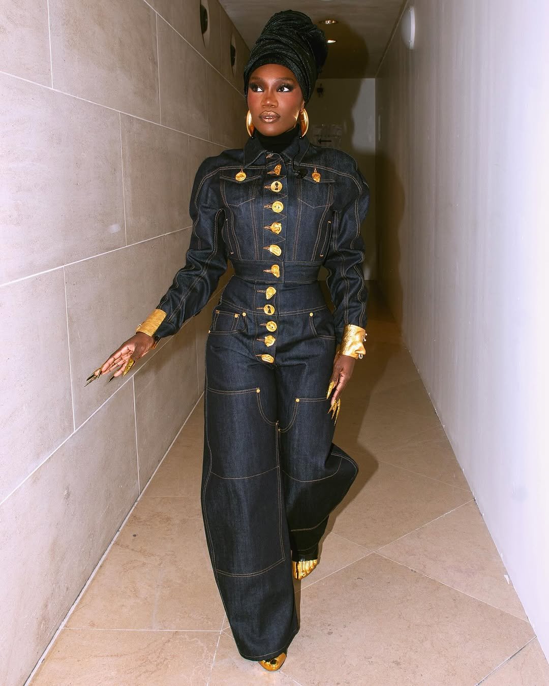 Pessoa em um elegante conjunto jeans escuro, com detalhes dourados e turbante combinando. Looks da Doechii