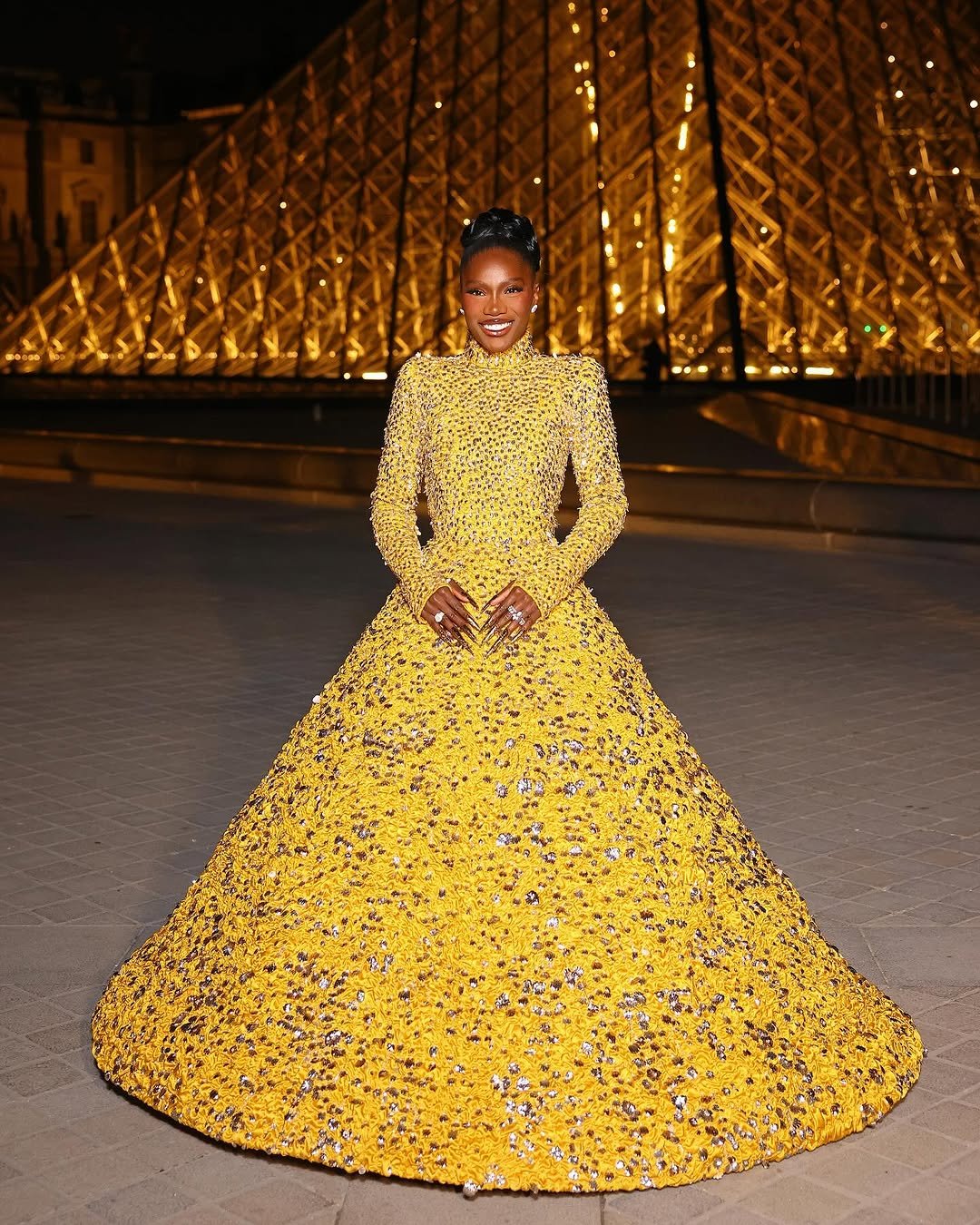 Looks da Doechii. Ela está usando um vestido amarelo bordado, com estrutura luminosa ao fundo.