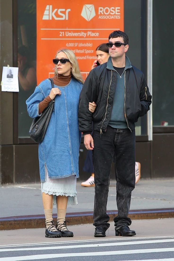 Um casal com roupas casuais caminha na rua. Ela usa vestido jeans e óculos escuros. Fachada ao fundo.