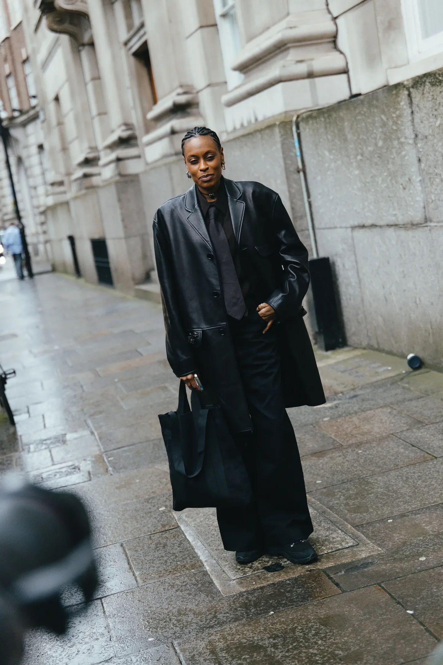 Pessoa em traje preto elegante, incluindo casaco longo, carregando bolsa como acessório de moda em rua urbana.