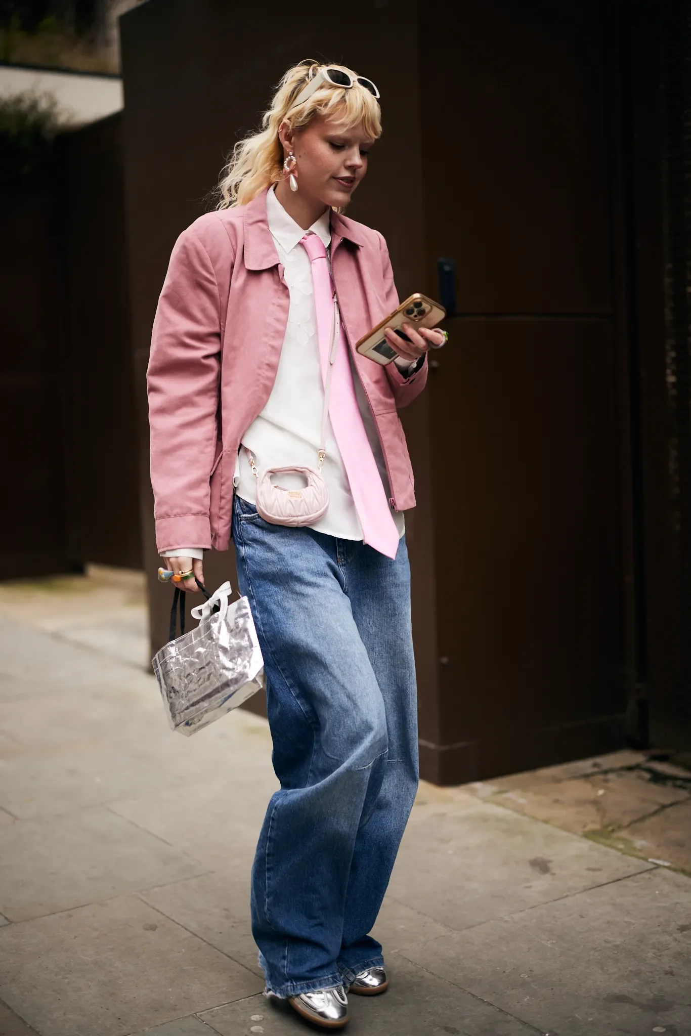 Mulher com jaqueta rosa e calça jeans larga, carregando bolsa prateada, olhando celular. Use de acessórios de moda.