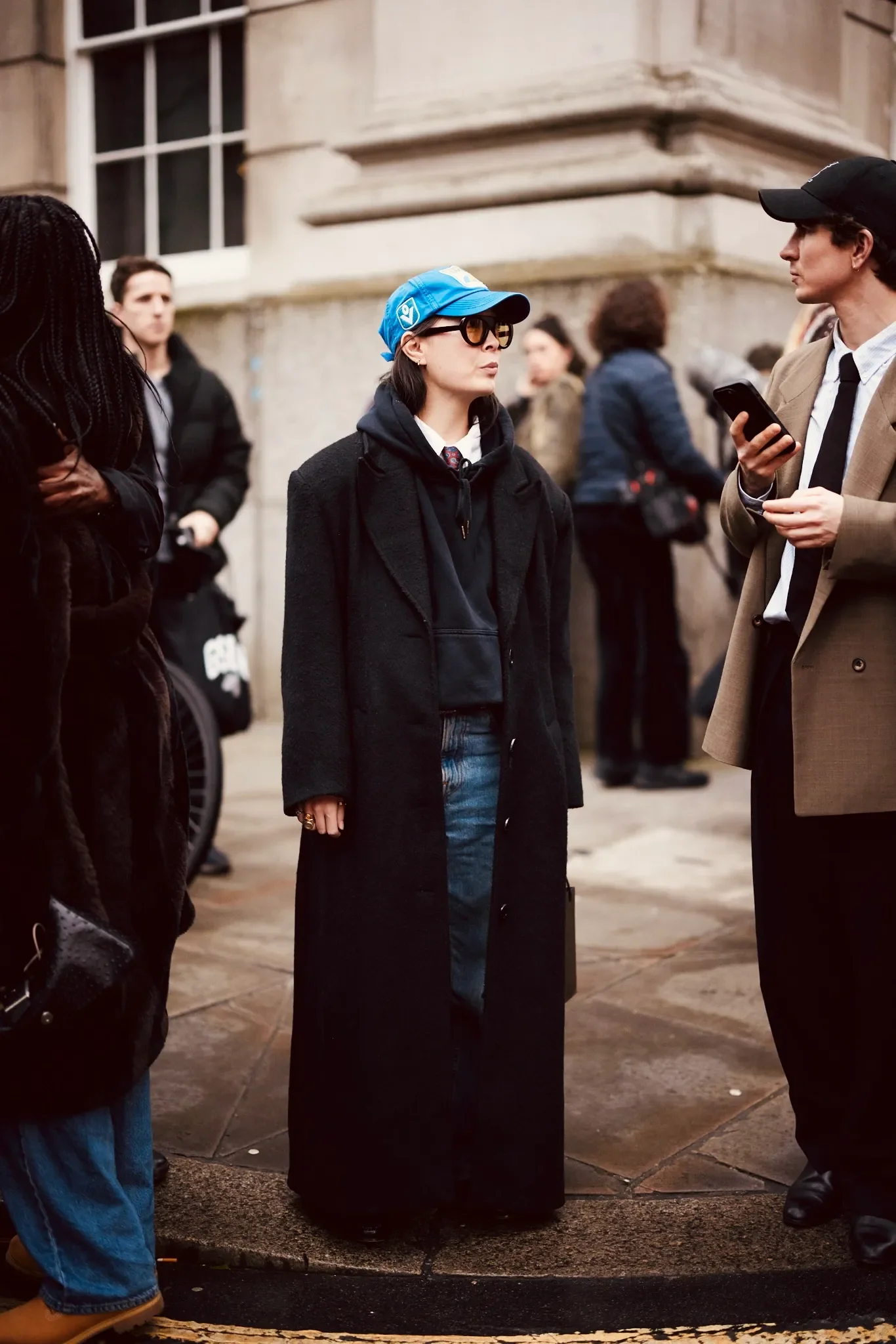 Pessoa usando boné azul como acessório de moda, óculos escuros, casaco longo preto e calça jeans.