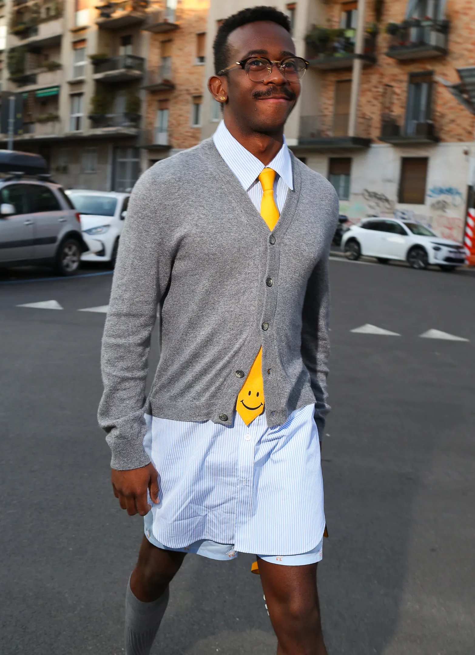Homem sorridente usa cardigan cinza com gravata amarela e camisa listrada, um moderno acessório de moda.