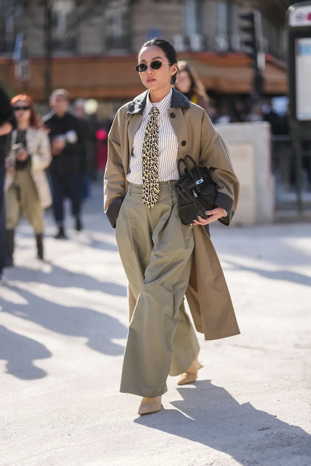 Pessoa usando trench coat bege, calças largas, gravata estampada e óculos de sol. Bolsa preta como acessório de moda.
