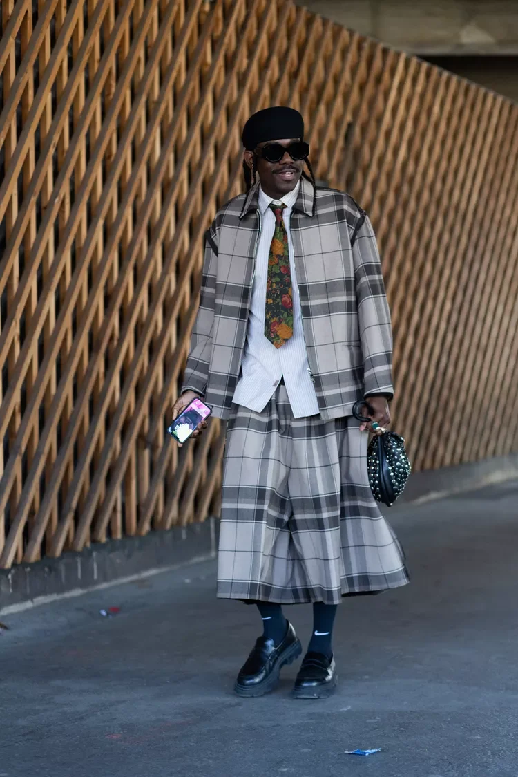 Pessoa usando um conjunto xadrez bege e cinza, gravata floral e chapéu preto, enquanto segura um acessório de moda.