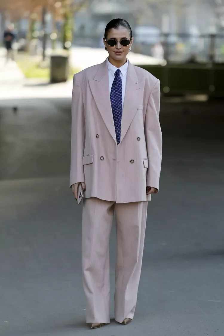Pessoa usando terno bege oversized, gravata azul e óculos escuros, exibindo estilo contemporâneo e acessório de moda.