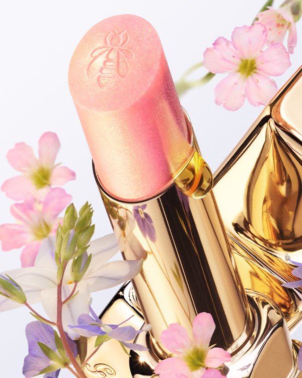 Batom rosa brilhante em embalagem dourada, cercado por flores delicadas, refletindo a sofisticação do Bee Beauty Secrets.