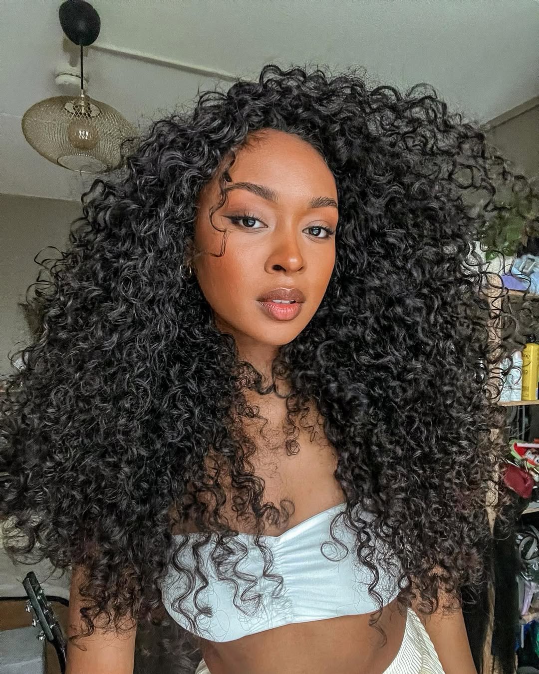 Cabelos cacheados volumosos destacam-se, sugerindo hidratações para cabelo cacheado. Mulher usa top branco.