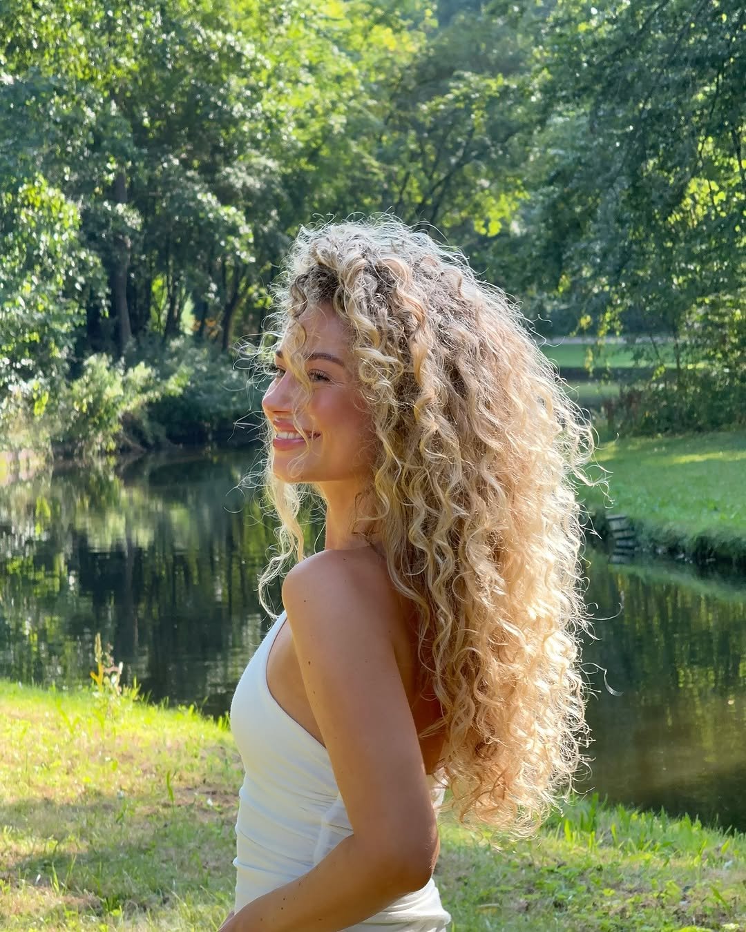 Pessoa sorridente com cabelo cacheado volumoso hidratações para cabelo cacheado em ambiente natural, próximo a um lago e rodeada por árvores.