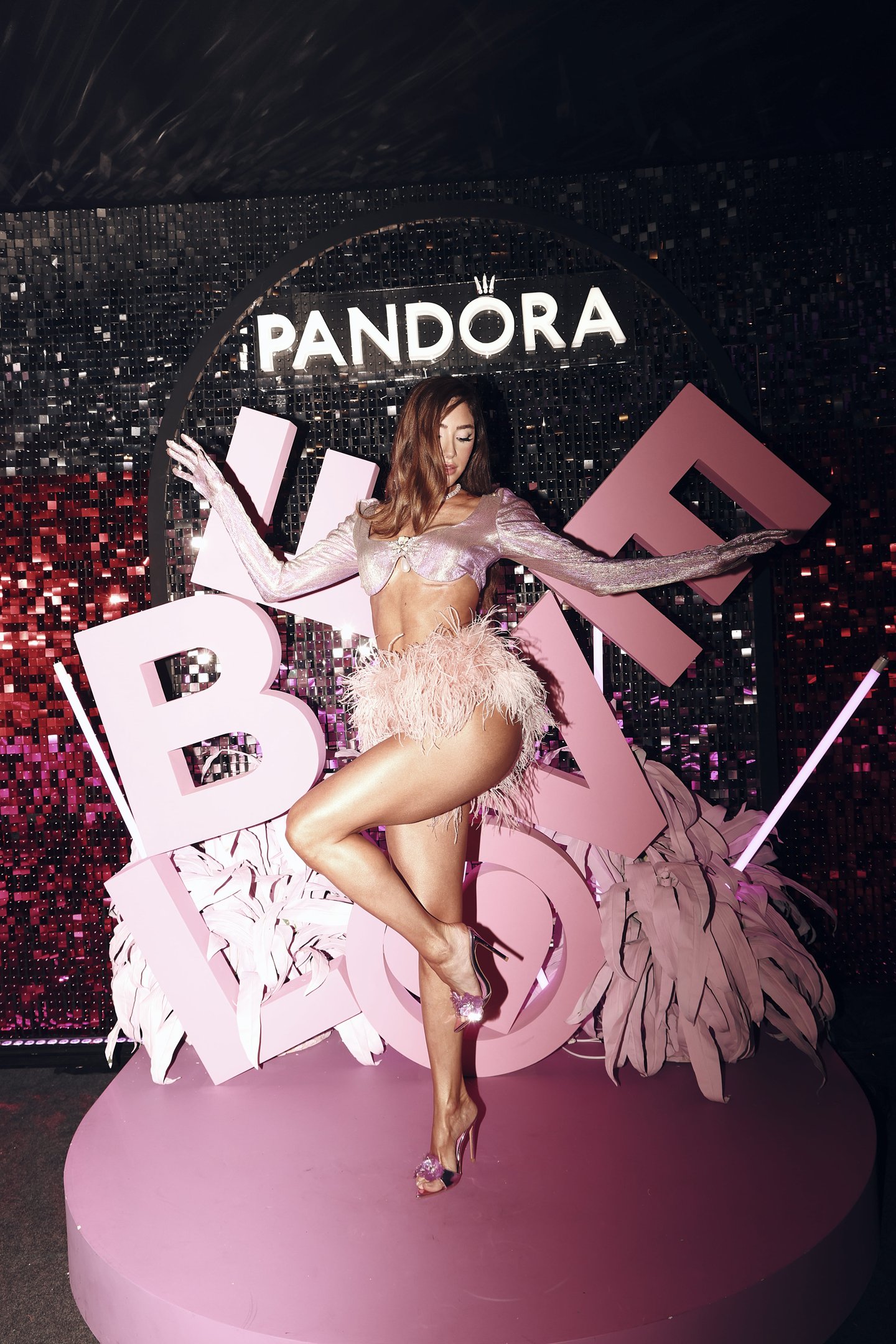 Mulher em pose de dança com traje rosa de plumas em cenário decorado com letras grandes e logotipo da Pandora.