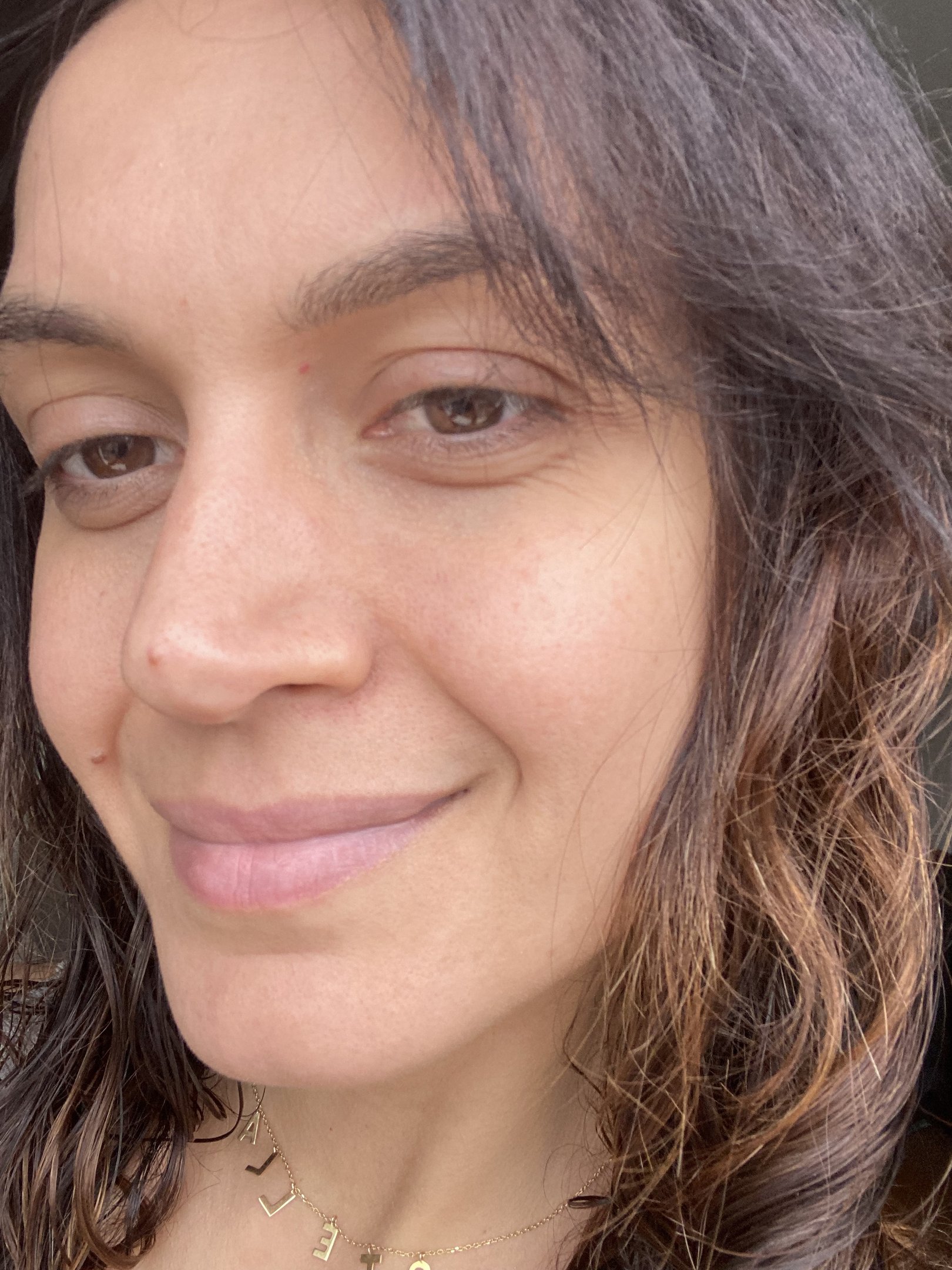 Pessoa sorrindo levemente, com cabelo castanho e ondulado, usando um colar dourado. Fundo neutro. após usar máscara de argila da benefit