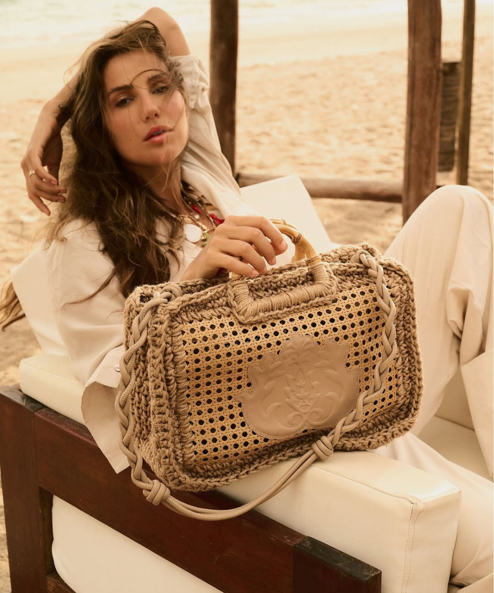 Mulher relaxando na praia com bolsa de palha detalhada nas mãos, sugerindo um estilo de moda ISLA.